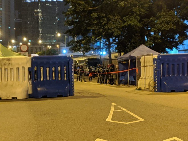 As dozens of people gather opposite the Chief Executive's Office on Tim Wa Avenue, riot police officers can be seen stationed near the government building. A police van arrives at the site and more officers emerge.